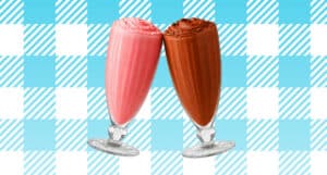 Two milkshakes in classic diner glasses lean together as if toasting. One is strawberry, and the other is chocolate. The shakes are set against a blue and white checkered background with diagonal stripe accents.