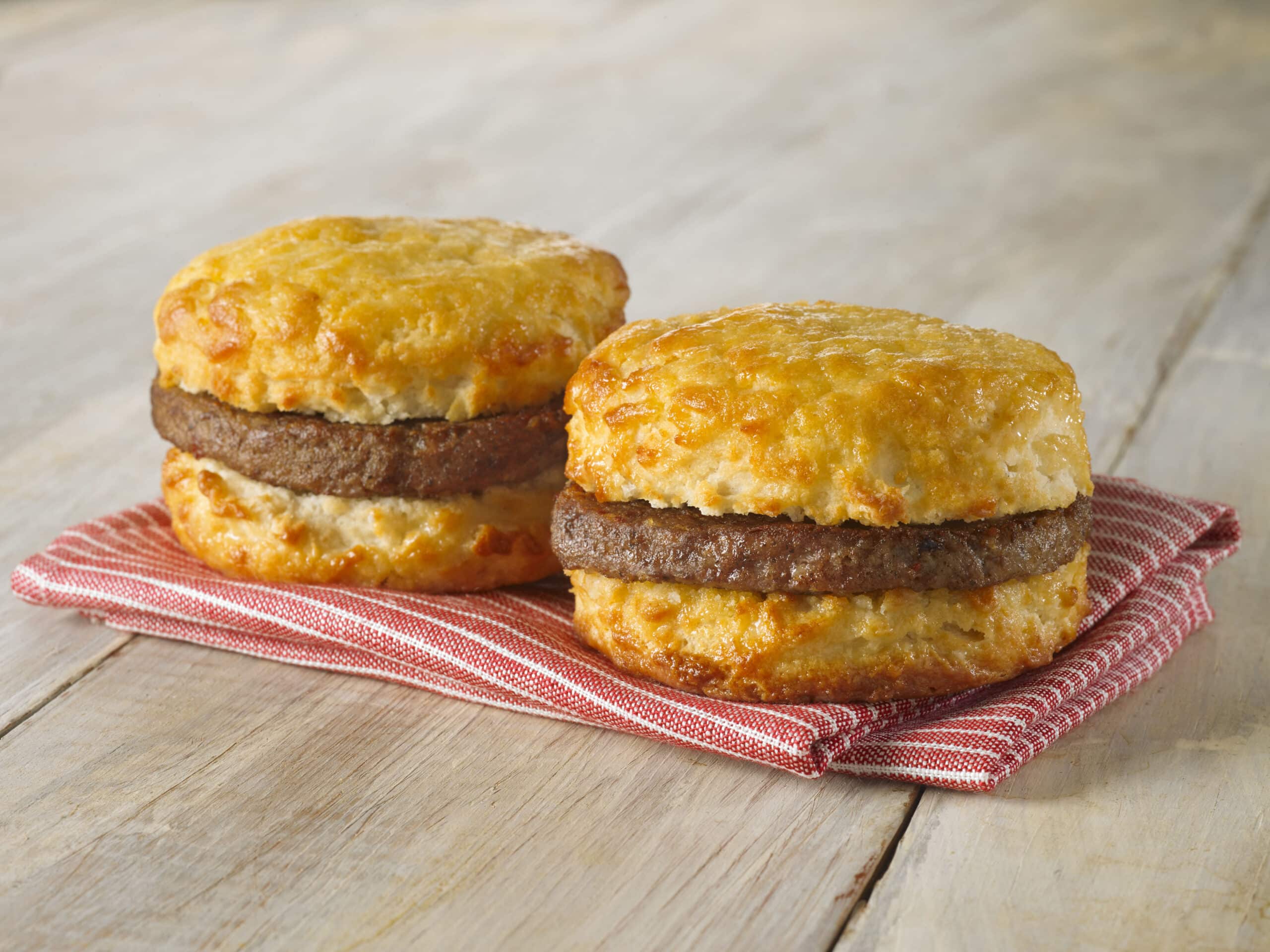 Two freshly baked sausage biscuits on a red and white checkered napkin, with golden brown biscuits sandwiching sausage patties.